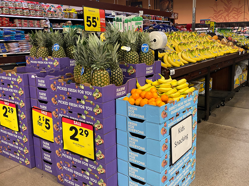 A row display in a store produce department. Boxes stacked in front include pineapples, with stacked boxes displaying oranges and bananas. Back rows include bananas and more fruit that is slightly unclear to the viewer.