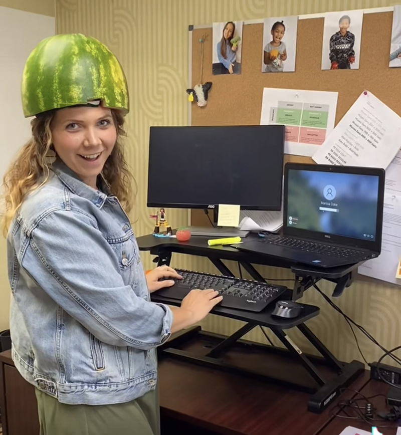 A woman works at her stand up desk with a watermelon on her head.