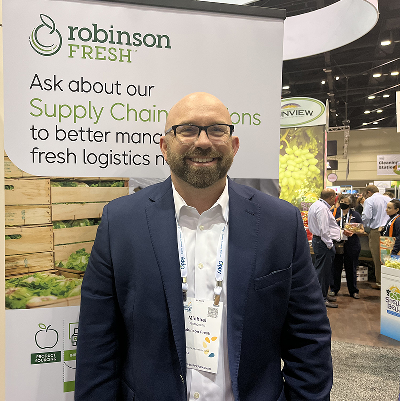 A man with glasses stands smiling in front of a Robinson Fresh ad. 