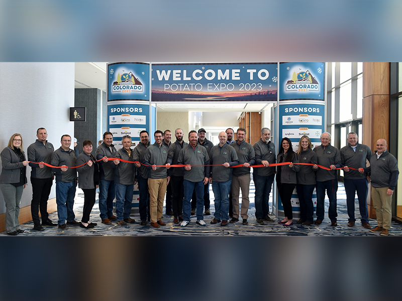 21 people stand side-by-side holding a red ribbon in a hallway, in front of a sign that says "Welcome to Potato Expo 2023."