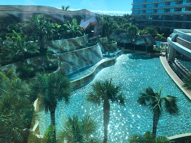 A view from above of an aqua blue pool, with hotel rooms to the right, a blue sky above, and palm trees in the foreground.